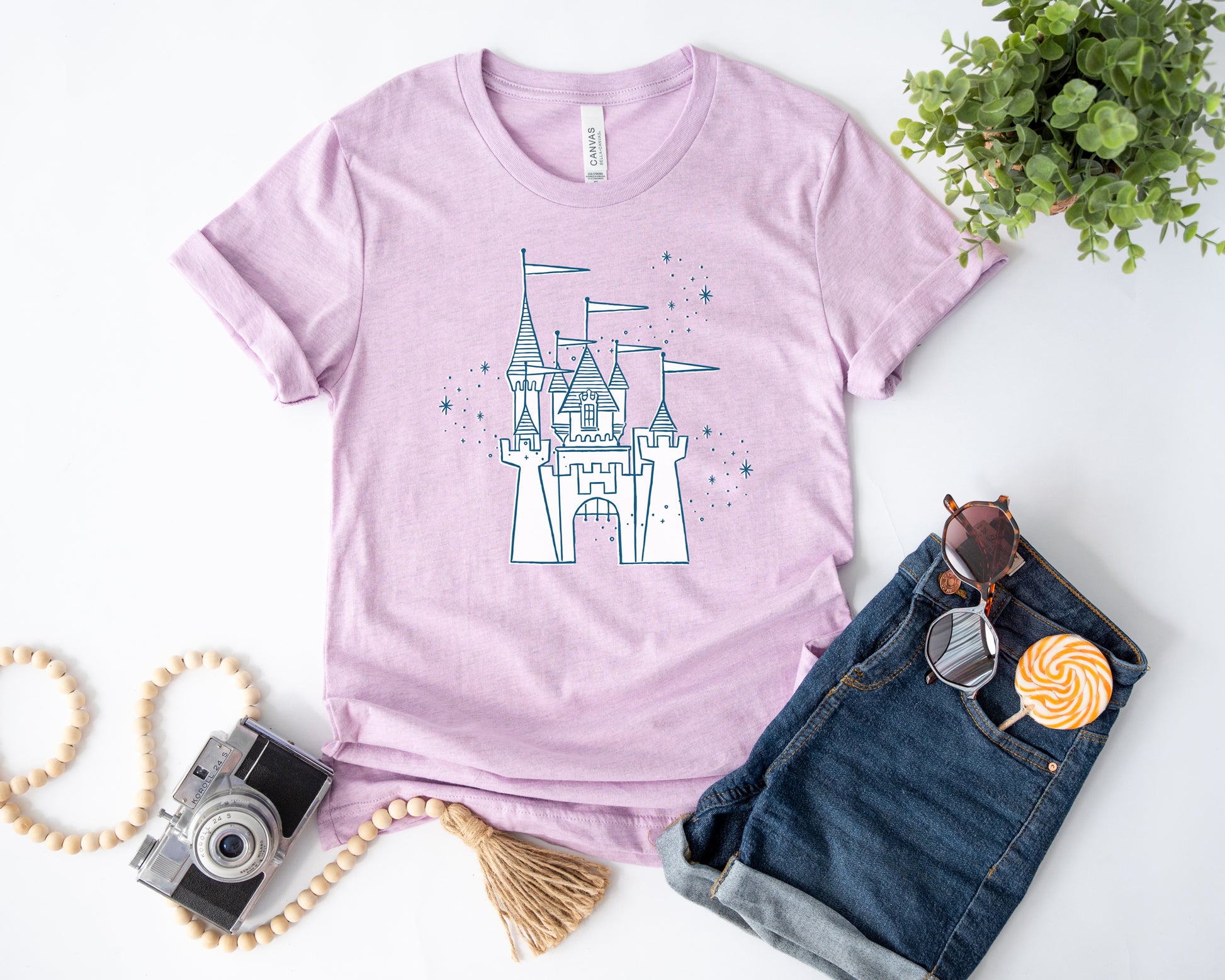 Flat lay of a purple shirt printed with vintage style sketch of the Disneyland Castle and pixie dust above the castle. The accessories next to the shirt are a camera, plant, shorts, sunglasses, and a lollipop.