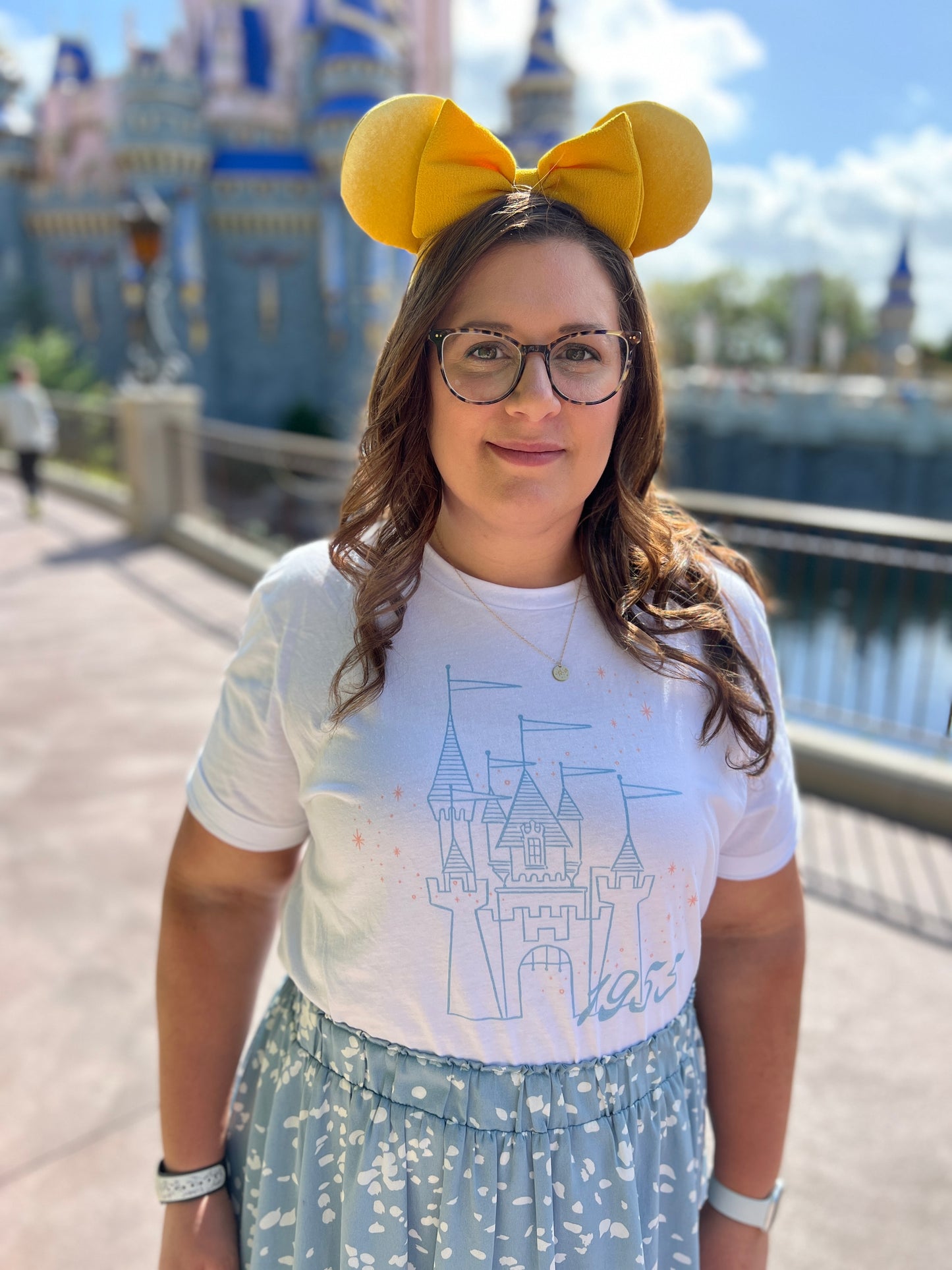 Woman in yellow mouse ears wearing a blue skirt and white t-shirt. The shirt is printed with vintage style sketch of a castle with the year 1955 and pixie dust above the castle.