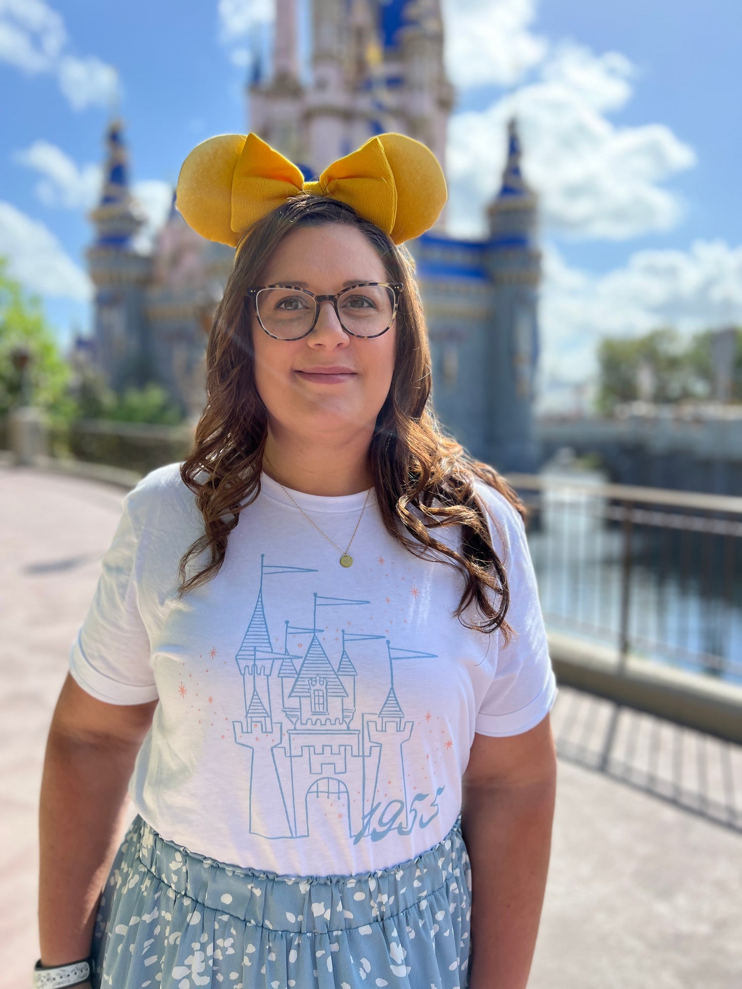 Woman in yellow mouse ears wearing a blue skirt and white t-shirt. The shirt is printed with vintage style sketch of a castle with the year 1955 and pixie dust above the castle.