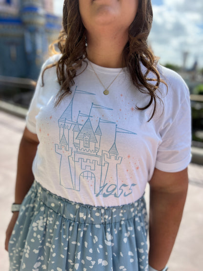 Woman wearing a blue skirt and white t-shirt. The shirt is printed with vintage style sketch of a castle with the year 1955 and pixie dust above the castle.