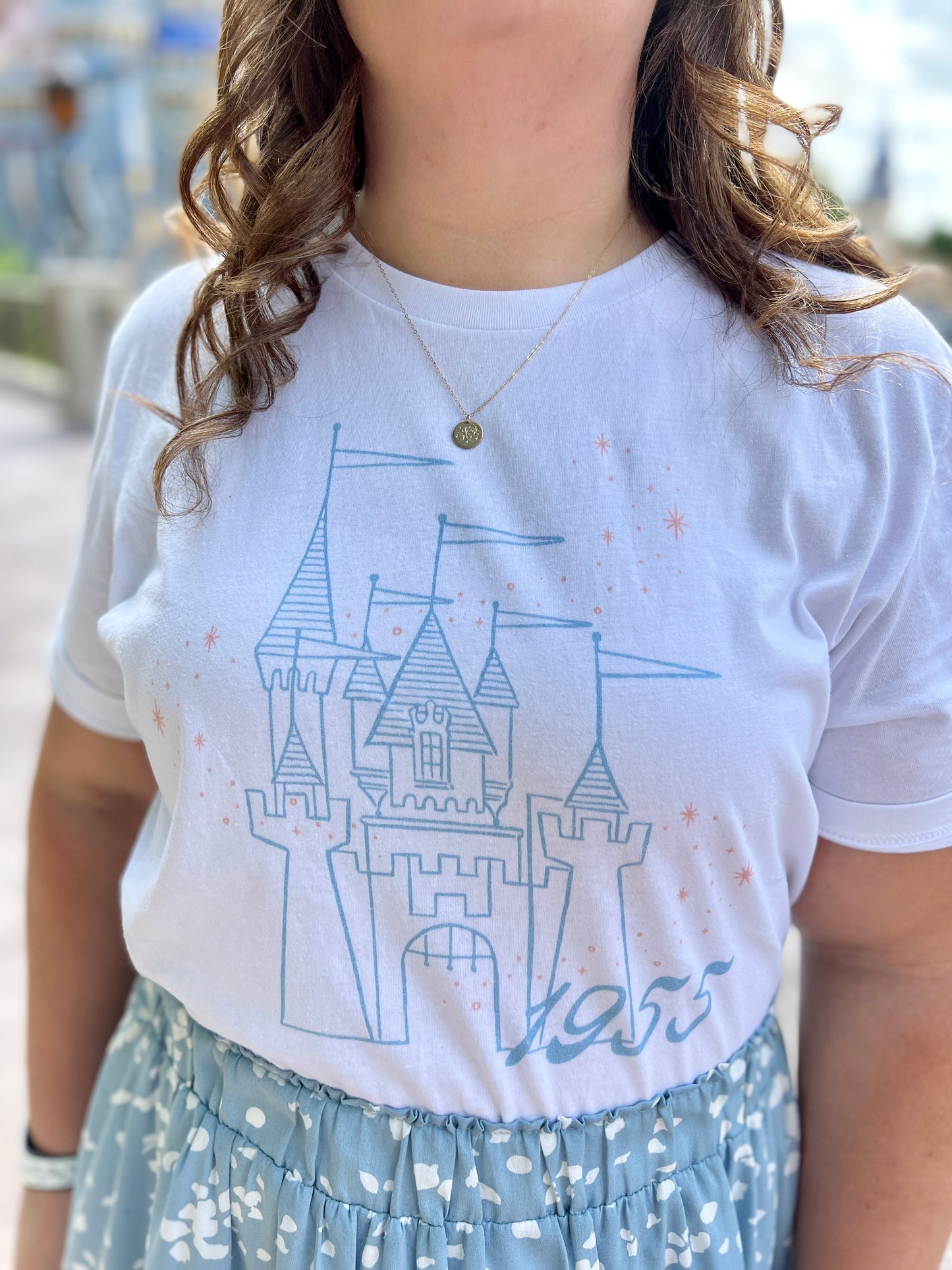 Woman  wearing a blue skirt and white t-shirt. The shirt is printed with vintage style sketch of a castle with the year 1955 and pixie dust above the castle.