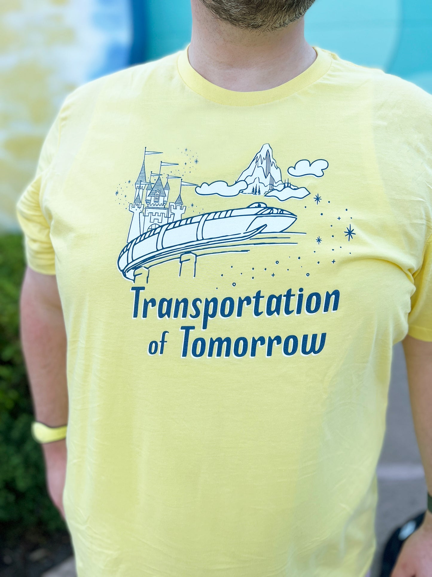 Man wearing a yellow shirt printed with vintage style sketch of the Matterhorn, Castle, and Monorail with pixie dust. The shirt says Transportation of Tomorrow.