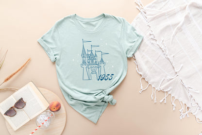 Dusty blue t-shirt printed with vintage style sketch of a with the year 1955 and pixie dust above the castle. There is a white blanket on the right side and a book, drink, glass, and peach in the bottom left corner.